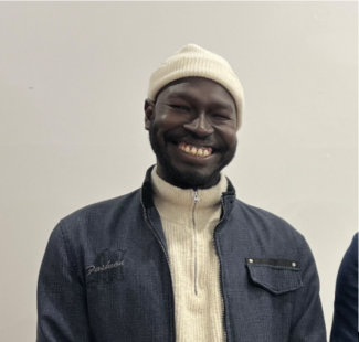 Ez Eldin Deng is a South Sudanese consultant. Ez is wearing a white ribbed beanie and cream ribbed swetaer with a denim jacket over the top. Ez is standing in front of a white wall with a wide smile