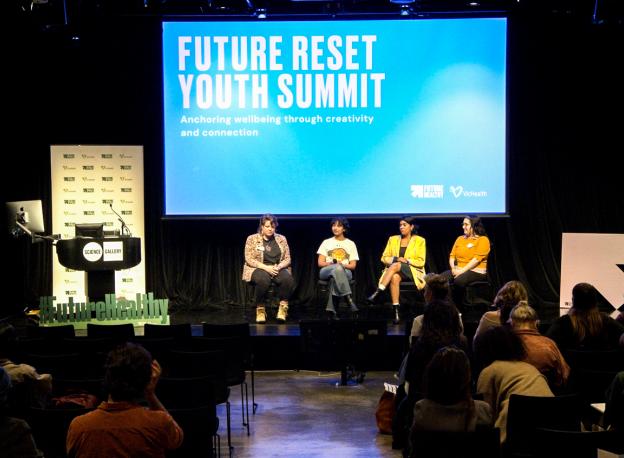 A panel of four is sitting on a stage. Behind them is a projector screen with the words "Future Reset Youth Summit" in bold and the words Anchoring Wellbeing through creativity and connection below.