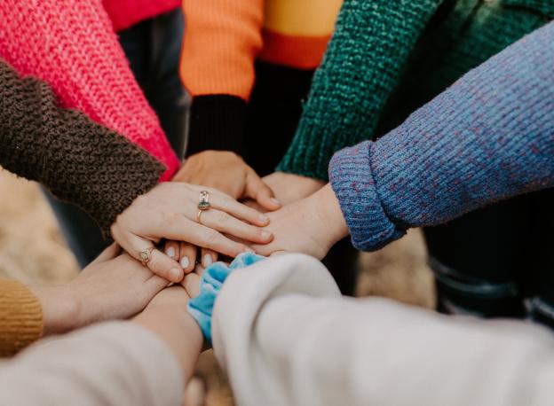 a group of people with lots of colourful knit jumpers on have put their hands all together in the spirit of teamwork. 