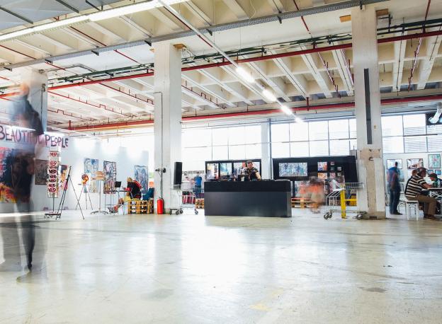 We see a big warehouse like space. The furniture is in the background and the foreground is bare apart form a blurred figure of a person possibly looking at an artwork out of frame. 