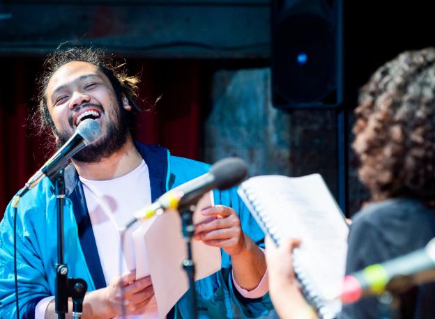 A man wearing a bright blue jacket is smiling while singing into a microphone. Another person is facing them we can see the back of their curly hair. They are both holding up notebooks as though this is a rehearsal. 